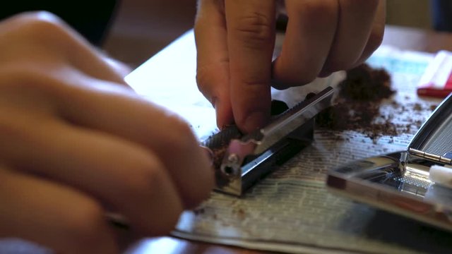 A young man makes a handmade cigarette filling paper with shredded tobacco using a rolling machine. On the table is a newspaper and an open cigarette case.