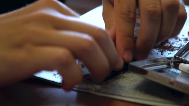 A young man makes a handmade cigarette filling paper with shredded tobacco using a rolling machine. On the table is a newspaper and an open cigarette case.