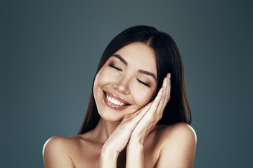 So cute. Beautiful young Asian woman keeping eyes closed and smiling while standing against grey background