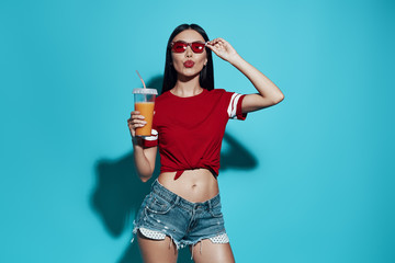 Juice cocktail. Attractive young Asian woman puckering and adjusting eyewear while standing against blue background