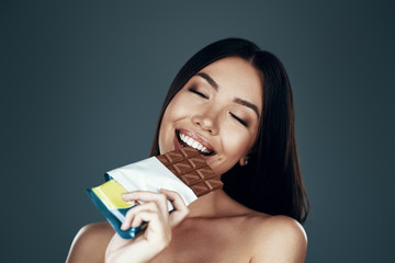 Real enjoyment. Beautiful young Asian woman smiling and eating chocolate while standing against grey background