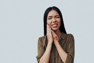 Terrible toothache. Frustrated young Asian woman making a face while standing against grey background