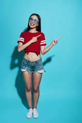 Look over there! Full length attractive young Asian woman smiling and pointing copy space while standing against blue background