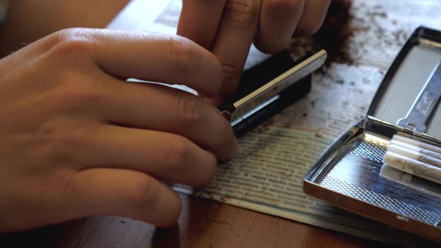 A young man makes a handmade cigarette filling paper with shredded tobacco using a rolling machine. On the table is a newspaper and an open cigarette case.
