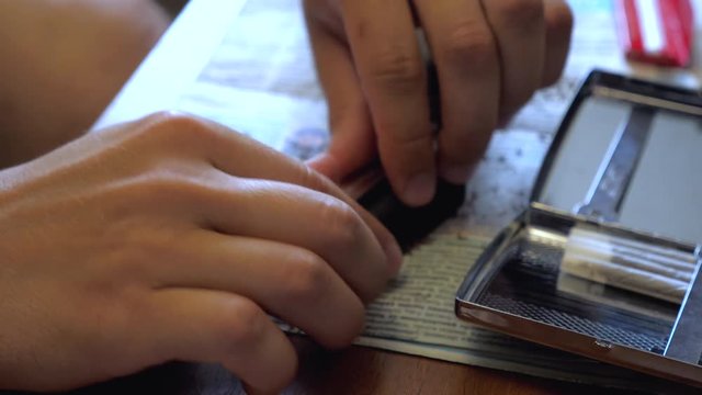 A young man makes a handmade cigarette filling paper with shredded tobacco using a rolling machine. On the table is a newspaper and an open cigarette case.