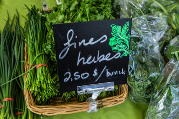 Organic produce sold at farmer's market. A close up view of a French sign showing fine herbs for sale on a market stall, bunches and bags of salad greens are seen behind, during a harvest fair.