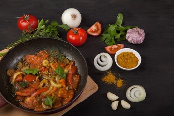 Roasted tomatoes with onions in a pan and fresh vegetables on a black background. Top view