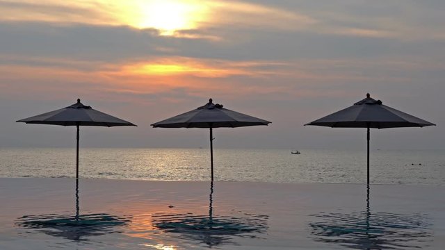 Parasole umbrellas reflecting in an infinity pool at a luxury beachfront location with the tropical setting sun behind over the ocean