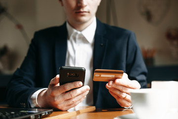Hand of a young businessman holding a gold credit card and a smartphone while sitting on a table indoor.