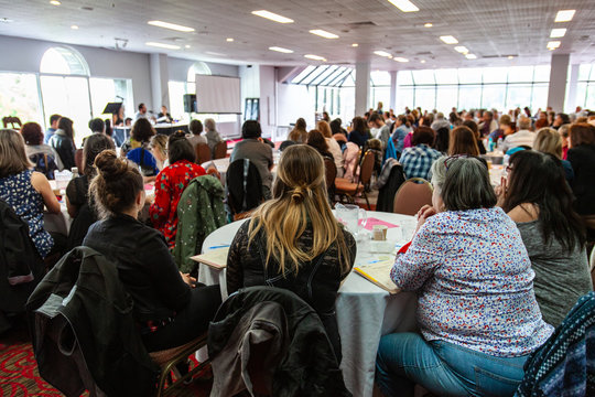 Atmosphere during corporate conference. A back view on a group of women sitting at a large table during a networking event for white collar workers. Lots of people can be seen watching a presentation