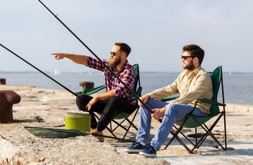 leisure and people concept - male friends with fishing rods sitting in camp-chairs on sea pier