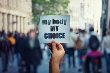 Feminist hands holding a protest banner with the message my body my choice over a crowded street. Human rights concept against fetus law and reproductive justice. Stop discrimination and injustice - obrazy, fototapety, plakaty