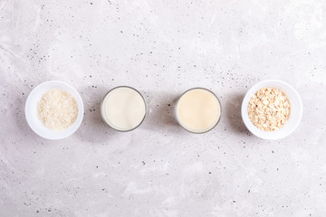 Glasses with rice and oat milk, bowls with rice seeds and oat flakes laid out in row on concrete background.
