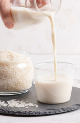 Female hand is pouring  rice milk from bottle into glass, and jar with rice seeds on light background. 