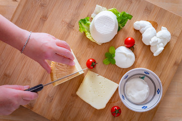 Female hands cutting a slice of pecorino cheese on a cutting board full of traditional Apulian cheeses: pecorino, mozzarella, ricotta, burrata. Puglia, Italy