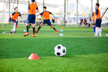 football on green artificial turf with blurry soccer team training, blurry kid soccer player jogging between marker cones and control ball with soccer equipment in soccer academy.