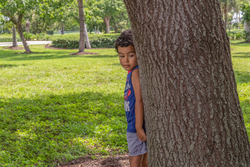 A small boy leans with his back on a large tree looking down. On a hot summer day, he enjoys playing under the tree shade in the park.