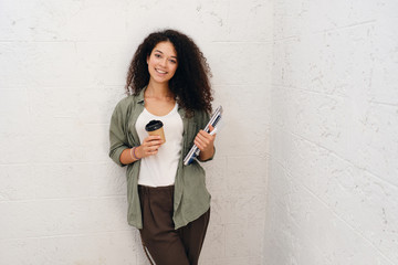 Young pretty smiling woman with dark curly hair in khaki shirt holding notepads and cup of coffee to go in hands happily looking in camera