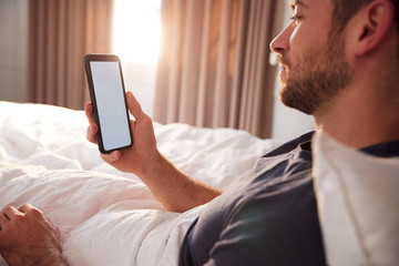 Man Sitting Up In Bed Looking At Mobile Phone After Having Woken Up