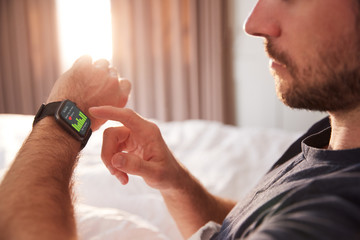 Man Sitting Up In Bed Looking At Screen Of Smart Watch