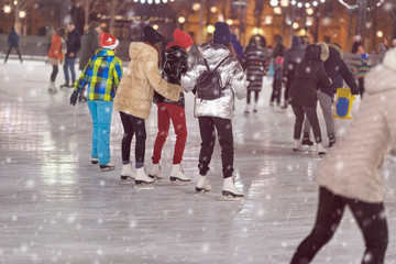 Group of girls skating back to us. Girlfriends ice skating in city park, snowy evening. Healthy outdoor winter activity