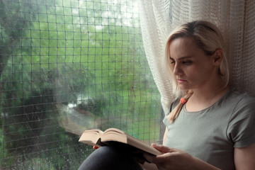 Blonde woman in casual clothes sitting by the window and reading book
