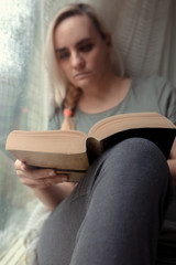 Blonde woman in casual clothes sitting by the window and reading a book