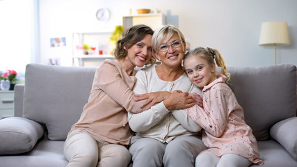 Multi-generation females hugging on home sofa smiling camera, family closeness