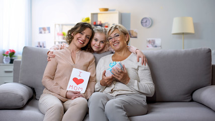 Cute girl hugging mother and granny with present box and hand-made greeting card