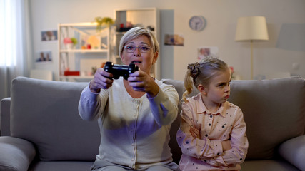 Grandmother playing video game with joystick, offended granddaughter sitting by