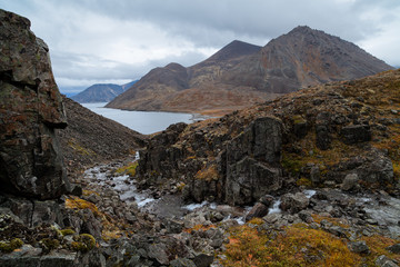 Severe northern landscape with a mountain stream in a gorge among the rocks. In the distance, a sea bay and mountains. Autumn in the Arctic. Providence Bay, Chukotka, Siberia, Far East of Russia.