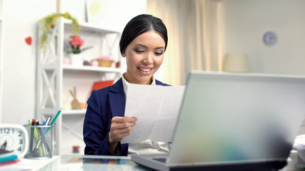 Young smiling female reading letter sitting front of laptop, correspondence