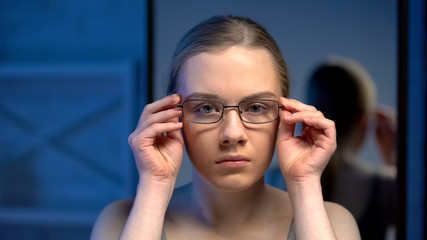 Unhappy young lady putting on eyeglasses, unsatisfied with mirror reflection