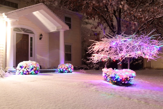 Christmas and New Year outdoor decoration background.Covered by fresh snow front yard of private house decorated for winter holidays.Night scene with glowing in the dark bushes, wrapped by garlands.