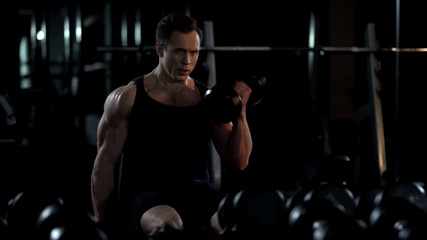 Handsome bodybuilder doing seated alternated biceps curl, training for summer