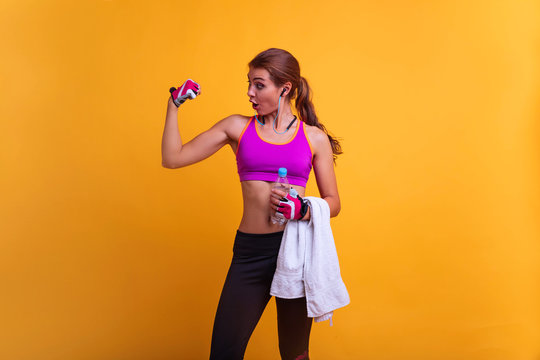 Image of a beautiful strong happy cheerful young sports woman posing and listening music with wireless earphones isolated indoors drinking water with towel on neck.