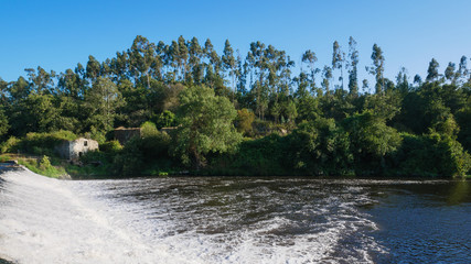 Wide angle view over Ave River in Touginho, Portugal on bright sunny summer day with blue sky. Fast flowing water over dam in foreground and trees on river bank.