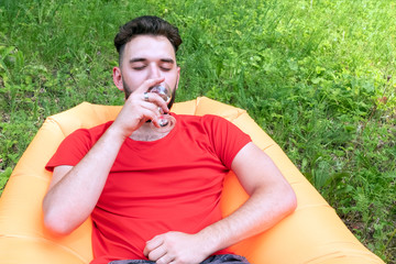 A guy with a beard in a red T-shirt is lying on an orange inflatable Lamzatz sofa. Caucasian traveler drinks wine at a picnic resting in the forest.