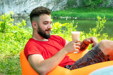 The guy with the beard in a red T-shirt lies on Lamzac orange inflatable sofa and holds a glass of beer in his hand. Caucasian traveler on vacation in the forest on the bank of a mountain river.
