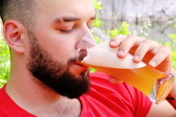 Close-up. Brutal guy with a beard in a red T-shirt drinks beer. Caucasian traveler on vacation in the woods uses alcohol. Portrait of a handsome young man.