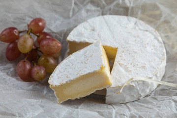Round cheese with a white mold is cut on pieces. On a section the crust and softness of structure is visible inside. It is decorated red grapes. Pergament sheet from below compositions