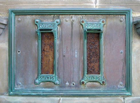 old british postal mail boxes with rusted letter slots and ornate green copper frames with the words postage stamps surrounded by an old stained metal frame in a stone wall