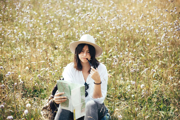 Hipster girl with wooden pipe looking at map and sitting in sunny  meadow, traveling in mountains....