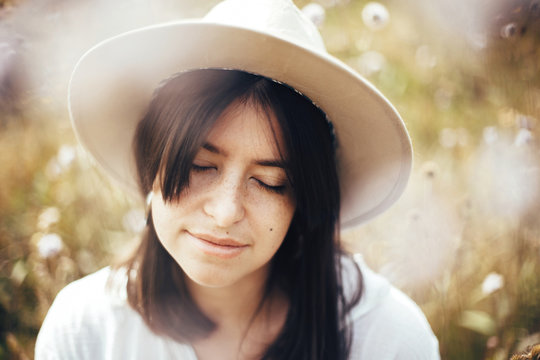 Portrait of calm hipster girl in hat relaxing in wildflower meadow,  traveling in sunny mountains. Stylish carefree woman enjoying hiking on top of mountain, sitting in herbs. Mindfulness