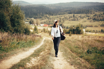 Hipster girl with backpack traveling on top of sunny mountain, walking on hills with woods. Stylish woman smiling enjoying hiking in mountains. Wanderlust and travel. Atmospheric moment