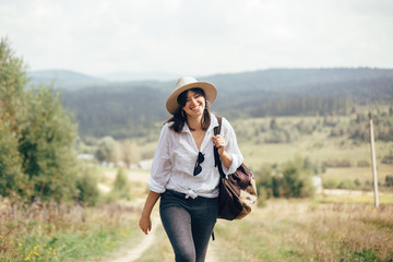 Happy hipster girl with backpack traveling on top of sunny mountain, walking on hills with woods. Stylish woman smiling and  enjoying hiking in mountains. Wanderlust and travel.