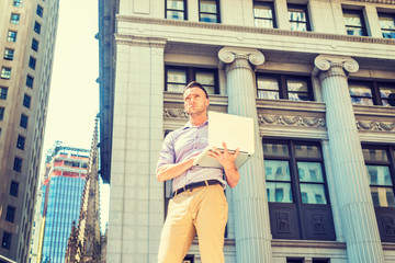 Young handsome man traveling, working in New York, wearing long sleeve shirt, beige pants, standing on street outside vintage office building, working on laptop computer, looking, thinking..