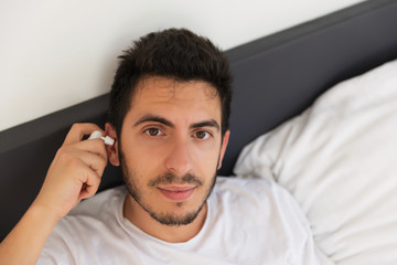 Obraz premium A young handsome man is sitting in his bed. He is holding a spray with medicine, he needs to cure his ear.