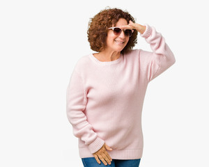 Beautiful middle ager senior woman wearing pink sweater and sunglasses over isolated background very happy and smiling looking far away with hand over head. Searching concept.