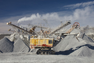 Excavator and large heaps of gravel on the background of smoke from the pipe of the quarry crusher in sunny day.  Mining industry. Quarry and mining equipment.